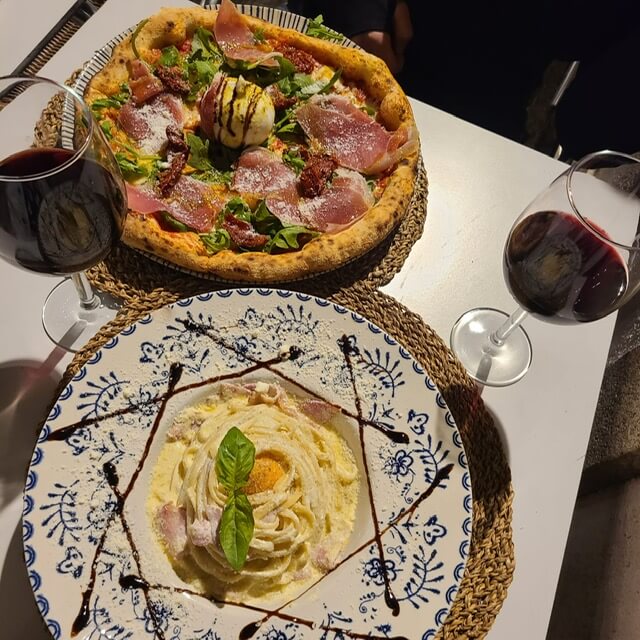 A table set with two glasses of wine, carbonara pasta, and pizza topped with meat, arugula, and burrata cheese