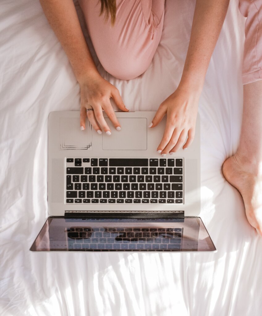 Chica sentada en la cama escribiendo en su computadora portátil