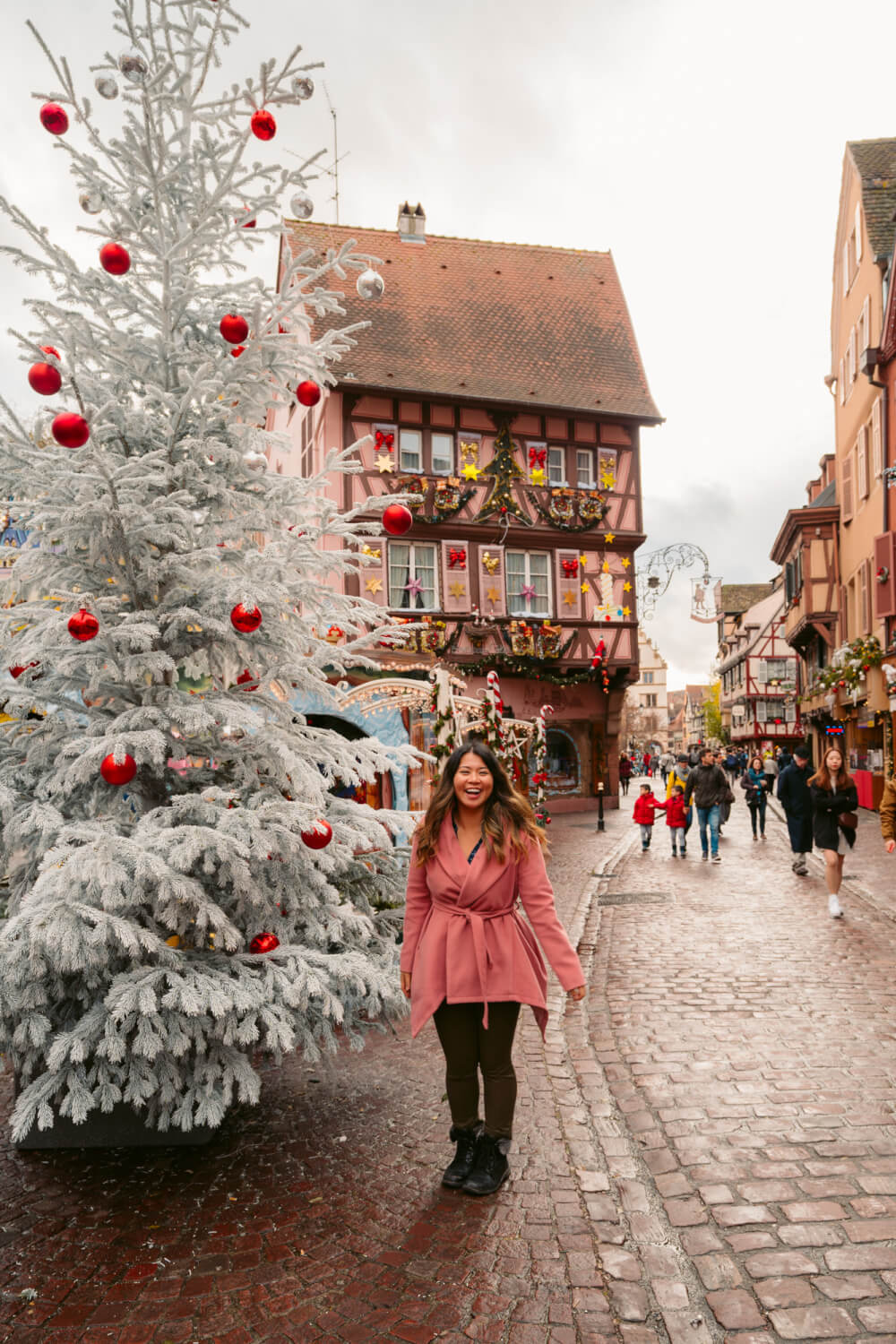 Travel blogger in pink coat posing in front of a white Christmas tree in Colmar, France