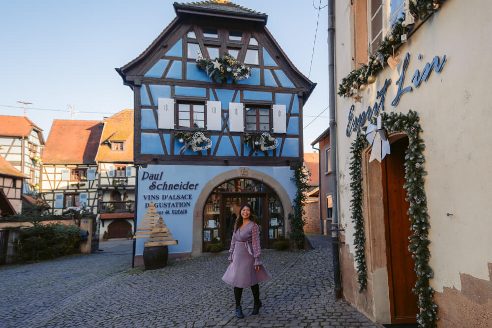 Travel blogger in purple dress in front of a blue house in Eguisheim