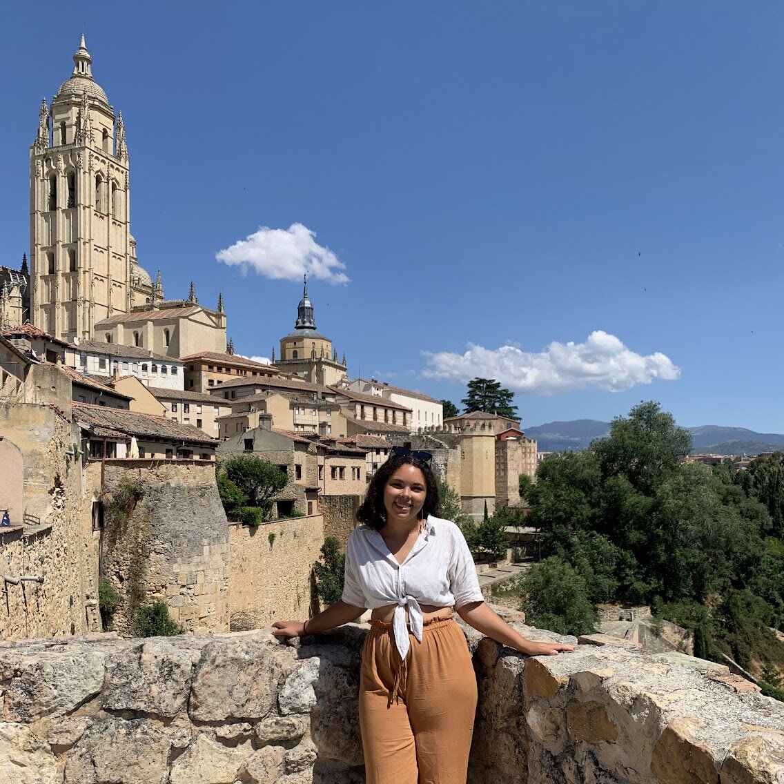 A person smiling leaning against a rock wall with a city in the background