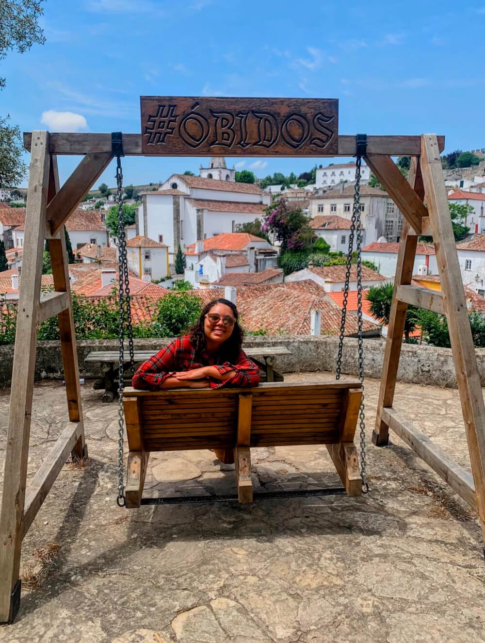 A person sitting on a swing under a sign that says "#Obidos"