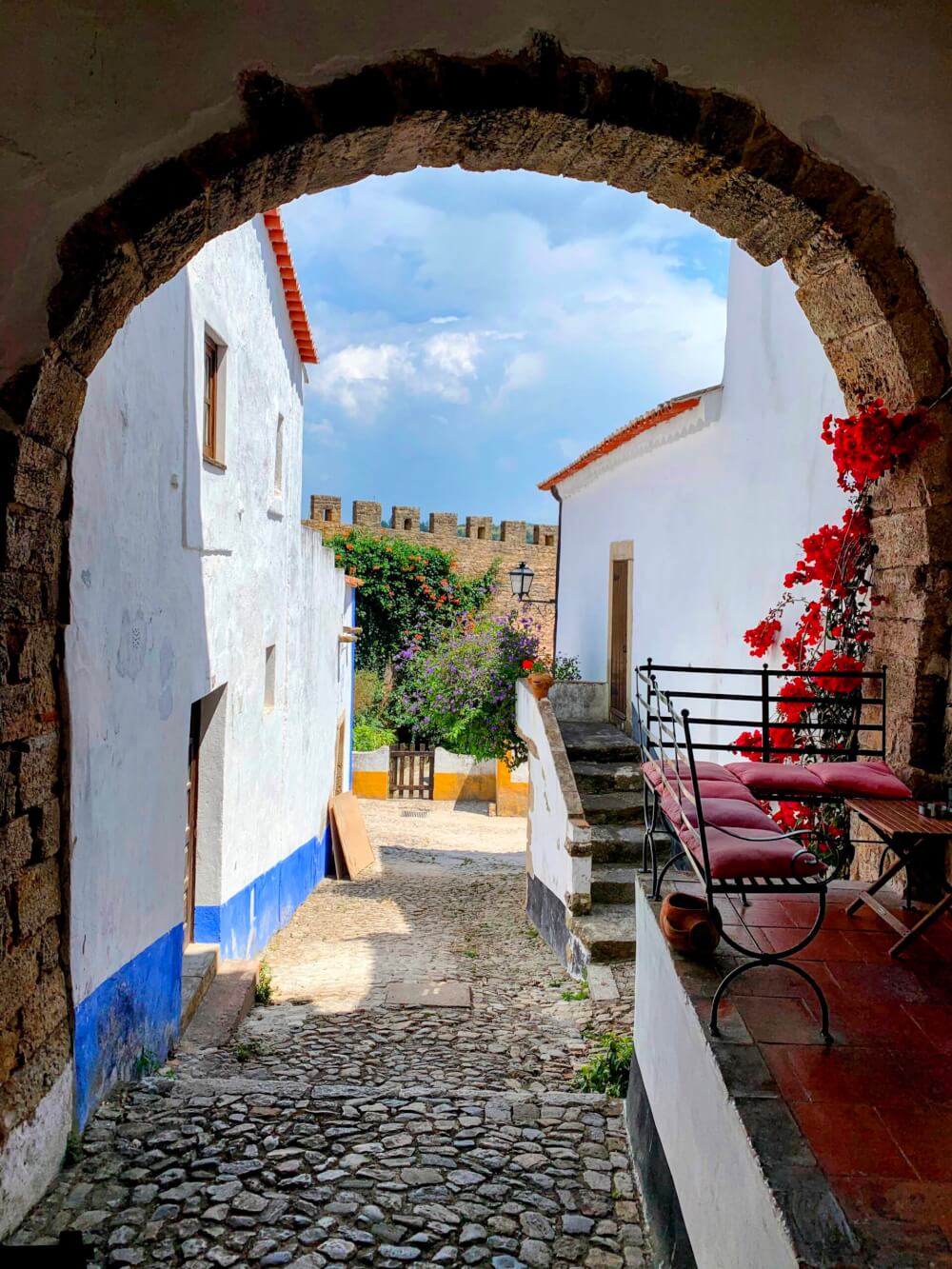 Outdoor restaurant seating under a stone archway, with flowers growing along the arch and a castle in the background