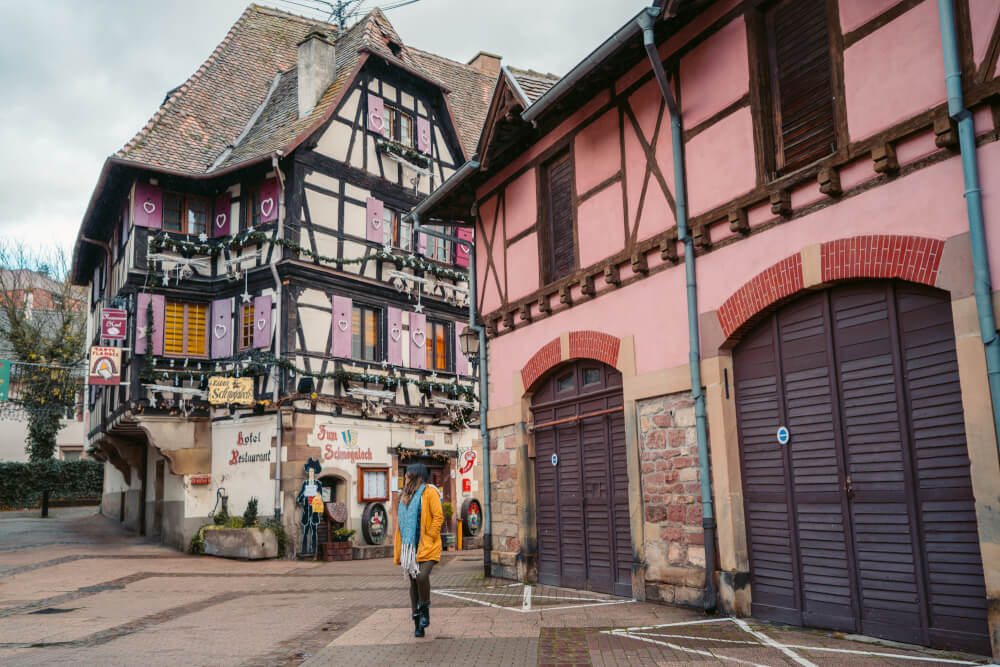 Travel blogger in yellow coat in front of a half timbered house with purple shutters in obernai