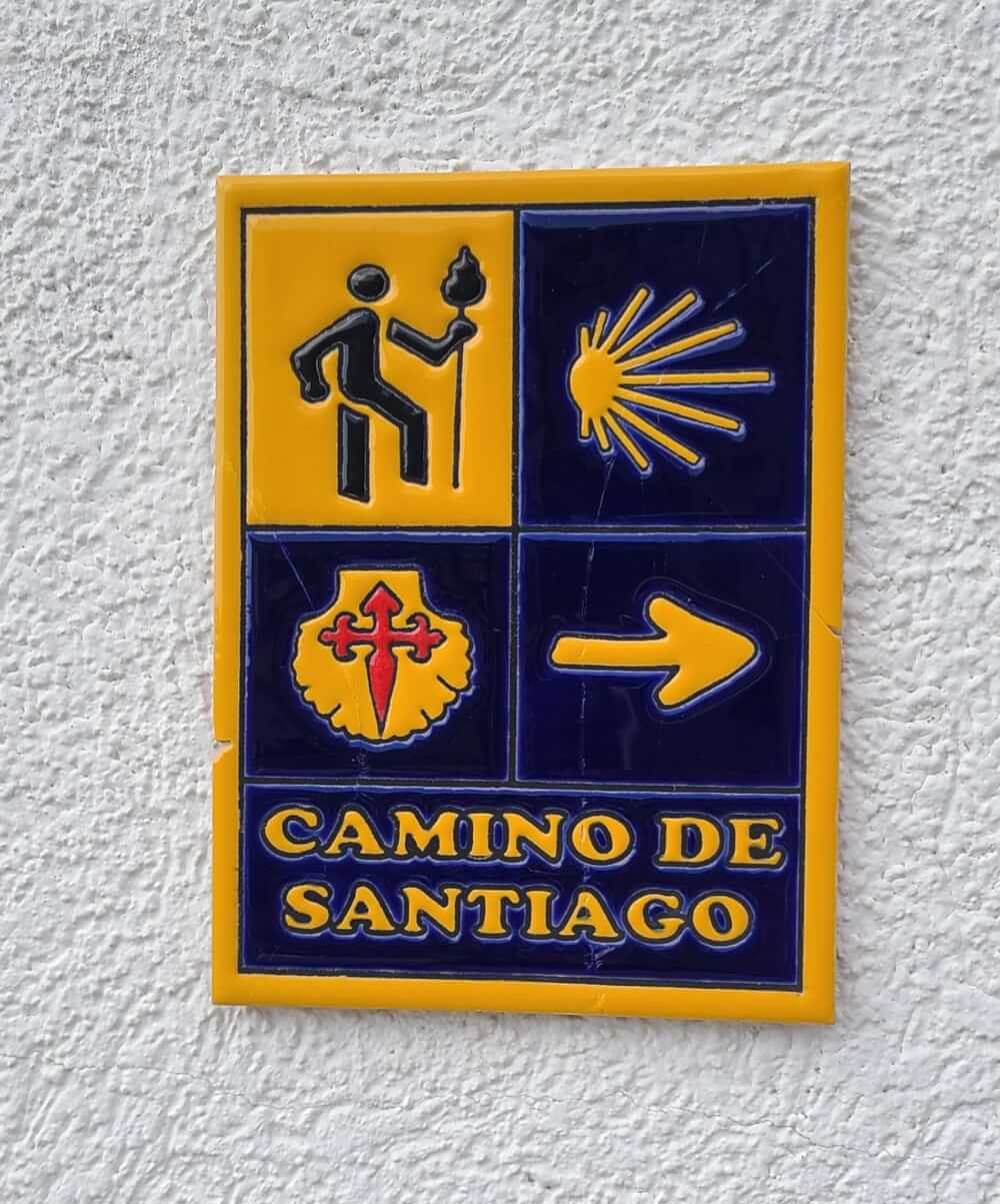 A blue and yellow sign that shows a person walking, an arrow, a scallop shell, and the words "Camino de Santiago"