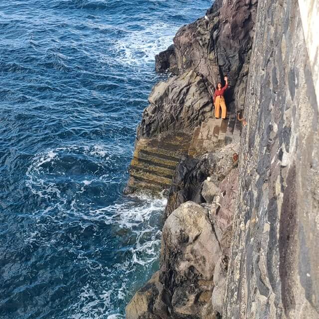 A woman waves at the camera from a staircase that goes down into the ocean