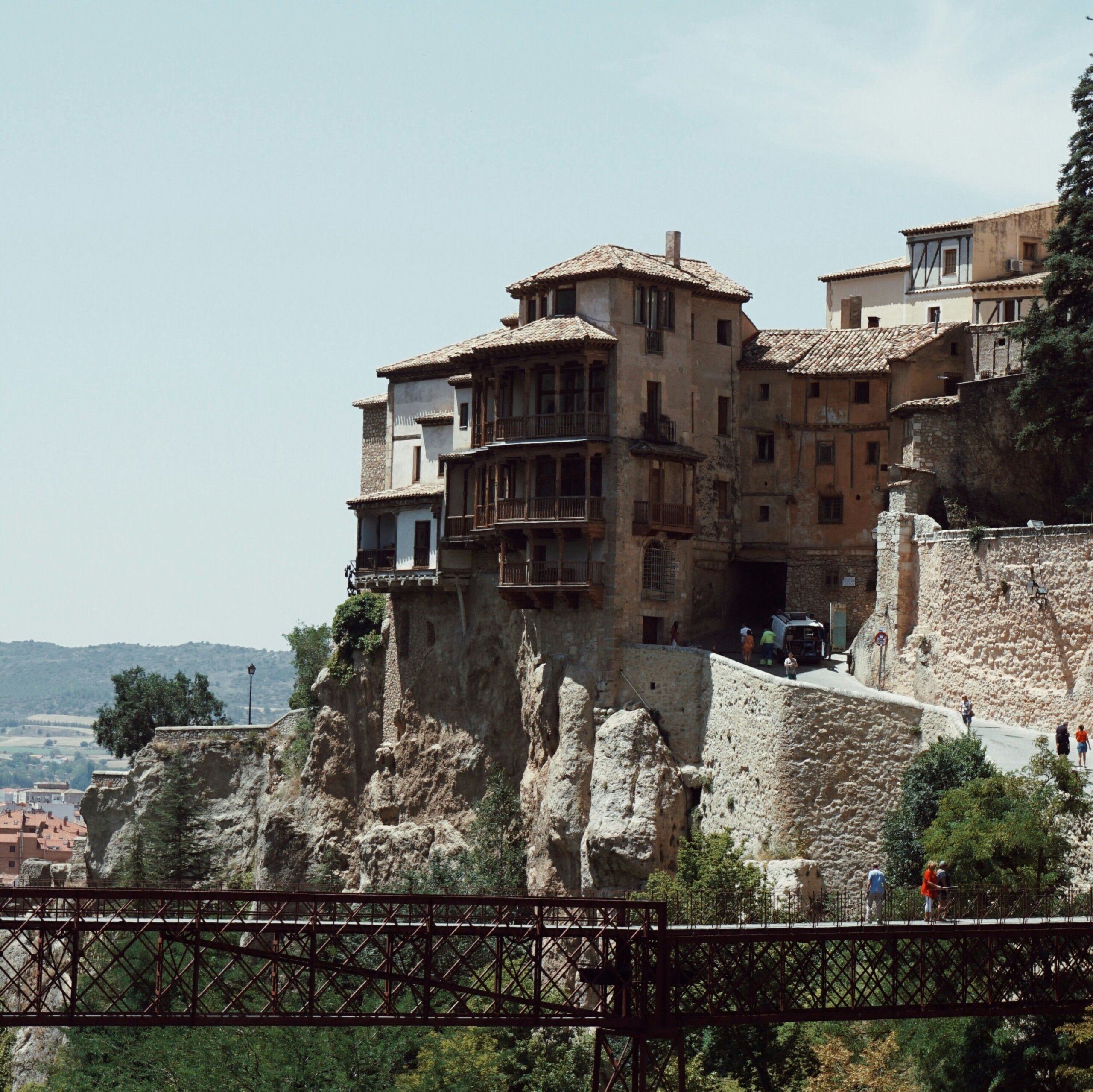 Two people standing on a metal bridge in front of a house built into a cliff in Cuenca, Spain