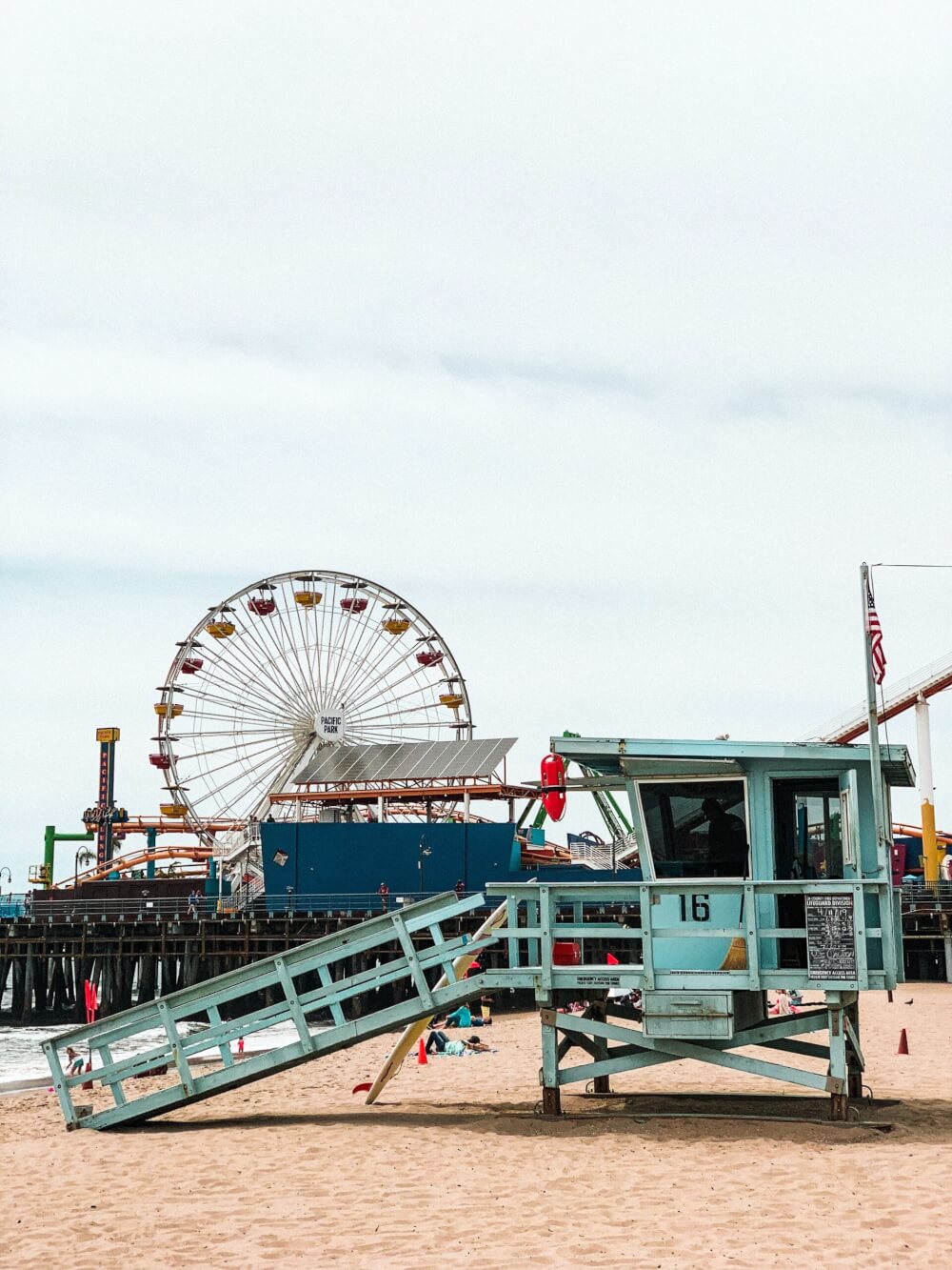 The pacific pier, with all its rides, the Ferris Wheel, and people laying on the beach. 