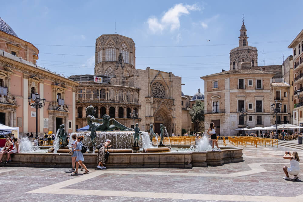 People in a European city square with a fountain, cafe, and cathedral. 