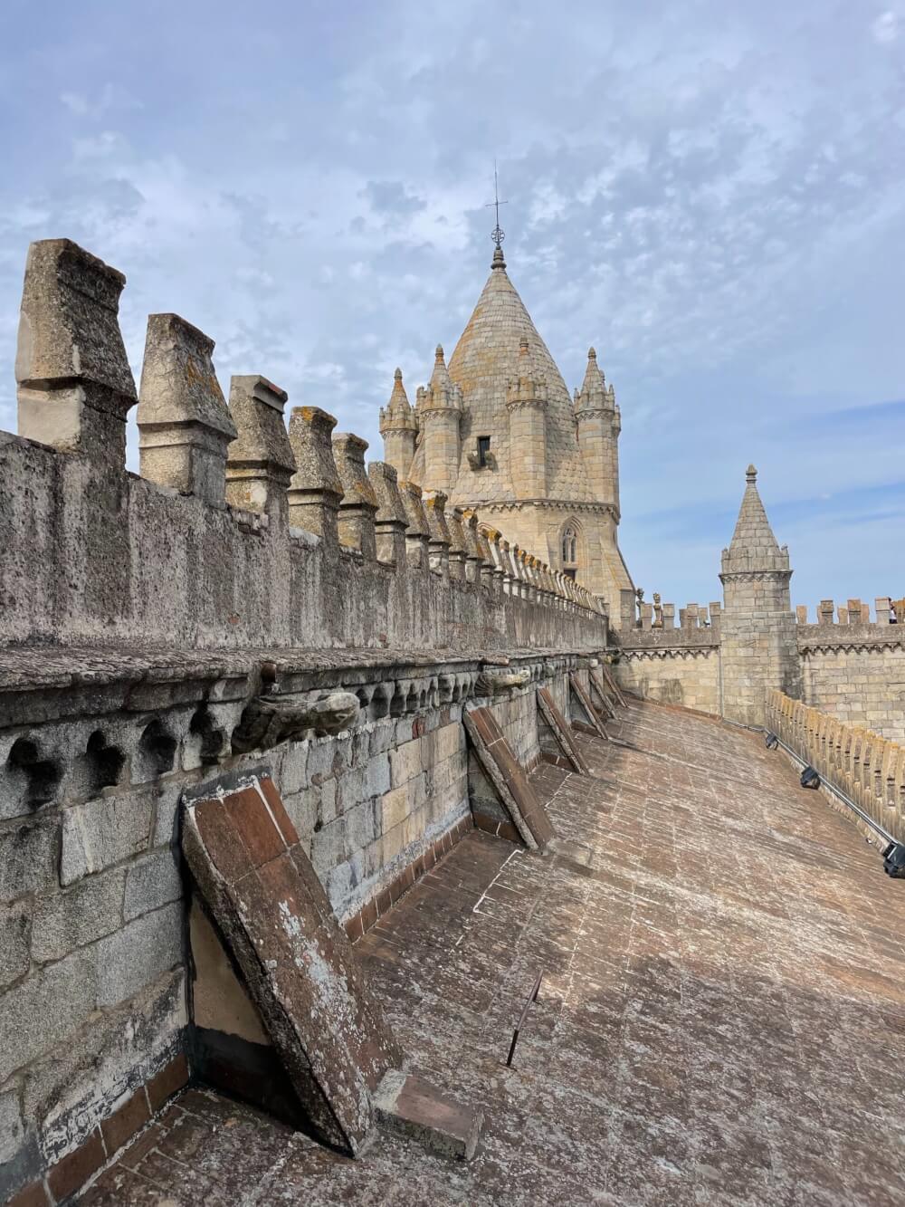 The roof of the cathedral of Evora. The picture shows a wall with stone spikes, a round medieval tower, and a sky full of clouds. 