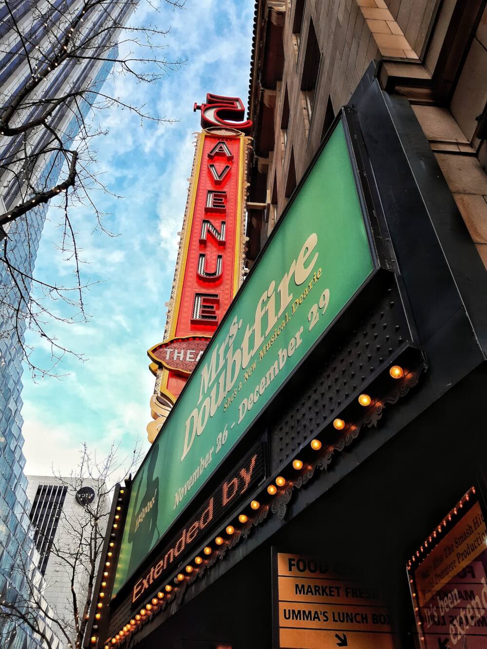 The marquis of a theater advertising Mrs. Doubtfire, with a blue sky in the background and a neon 'THEATER' sign. 