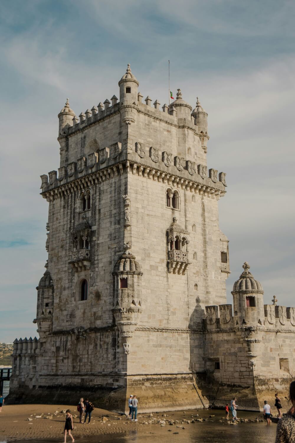 A grey tower on a beach with people walking along the shore