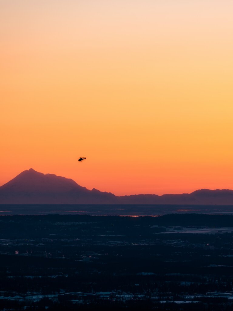 A view of a helicopter  flying over a mountain range, superimposed over a lurid sky.  