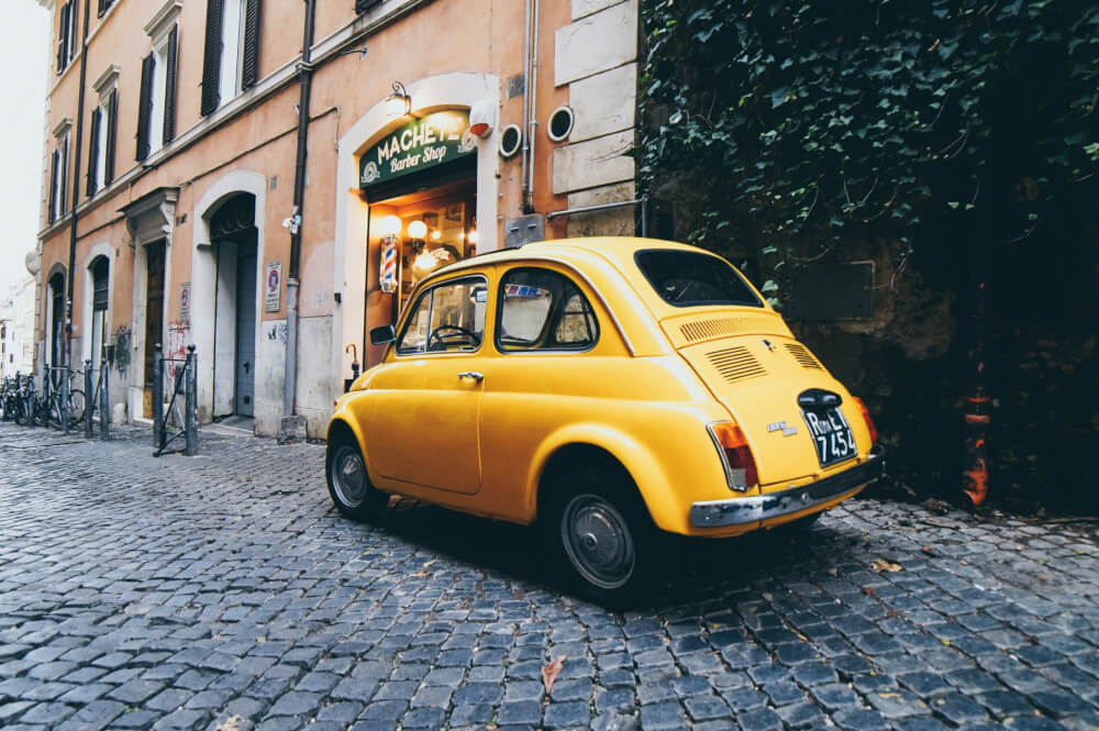 A yellow vintage Fiat parked on a cobbled street in Rome.